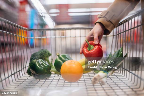 close-up of female hand placing fresh organic fruits and vegetables in shopping trolley - oranges in basket at food market stock pictures, royalty-free photos & images