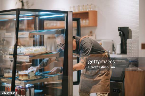 asian chinese senior man retrieving slice of cake dessert to serve to his customer from kitchen counter - boulangerie vitrine stockfoto's en -beelden