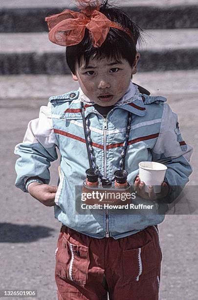 Portrait of an unidentified young girl, with a red bow in her hair, and snack cup in one hand, and a pair of binoculars around her neck, Beijing,...