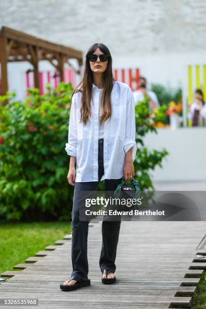 Leia Sfez wears sunglasses, a white t-shirt, a white oversized shirt, black denim jeans pants, a black leather mini Hermes Kelly bag with attached...