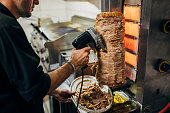In the kitchen of the Greek restaurant, the chef using the electric tool to cut the chicken gyros from the gyros grill