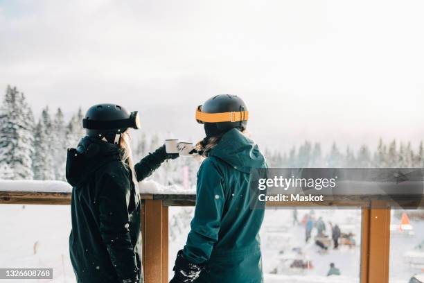 female friends toasting coffee cups while standing by railing during winter - vacaciones en la nieve fotografías e imágenes de stock