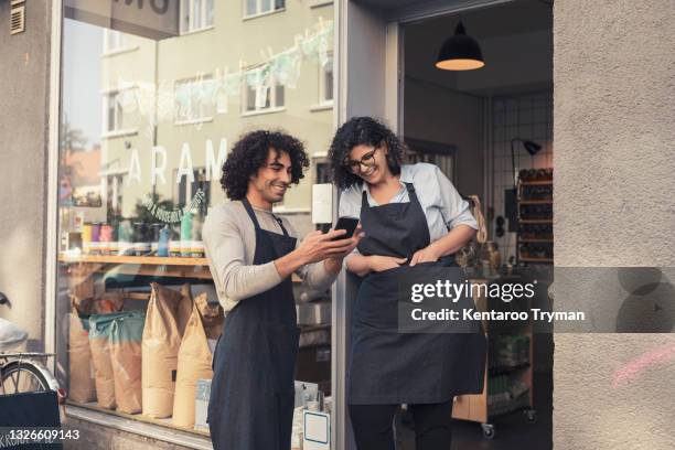 cheerful male and female owners sharing smart phone at doorway - happy merchant fotografías e imágenes de stock