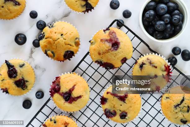 image of black, metal cooling rack containing batch of freshly baked blueberry muffins in paper cake cases besides bowl of fresh blueberries, on marble effect background, elevated view - muffin stockfoto's en -beelden