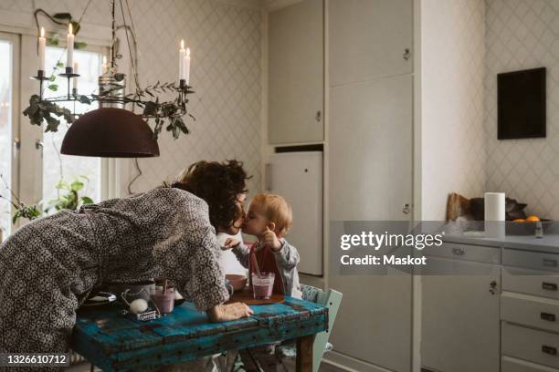 affectionate mother kissing daughter sitting at dining table in kitchen - västra götalands län stockfoto's en -beelden