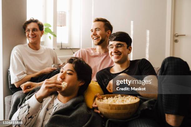 smiling male friends eating popcorn while watching sports at home - manly room stock pictures, royalty-free photos & images