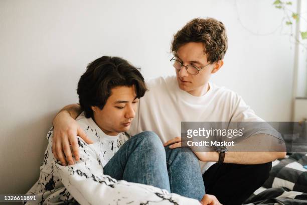young man sitting by worried male friend in bedroom - emotional support stock pictures, royalty-free photos & images