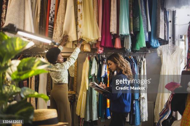 female owner making notes while colleague arranging clothes in boutique - fashion store staff stock pictures, royalty-free photos & images