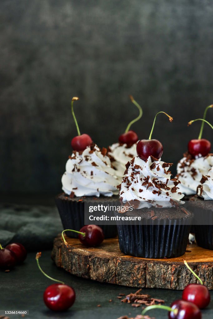 Image of batch of homemade, Black Forest gateau cupcakes in brown paper cake cases on wooden cake stand, piped whipped cream rosettes topped with morello cherries sprinkled with chocolate shavings, black background, focus on foreground