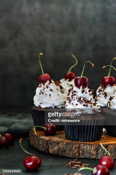 image of batch of homemade, black forest gateau cupcakes in brown paper cake cases on wooden cake stand, piped whipped cream rosettes topped with morello cherries sprinkled with chocolate shavings, black background, focus on foreground - forma de queque imagens e fotografias de stock
