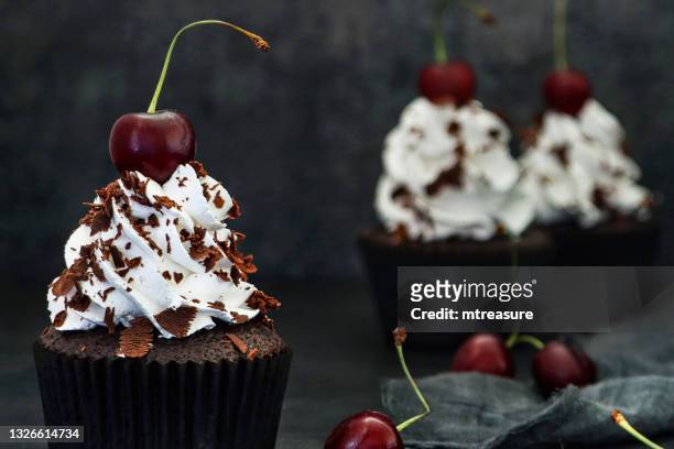 image of batch of homemade, black forest gateau cupcakes in brown paper cake cases, piped whipped cream rosettes topped with morello cherries sprinkled with chocolate shavings, beside grey tea towel against black background, focus on foreground - brown paper towel stock pictures, royalty-free photos & images