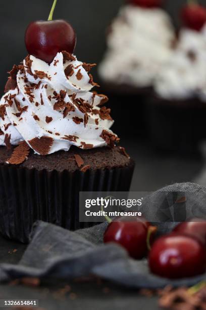 close-up image of batch of homemade, black forest gateau cupcakes in brown paper cake cases, piped whipped cream rosettes topped with morello cherries sprinkled with chocolate shavings, beside grey tea towel against black background, focus on foreground - brown paper towel stock pictures, royalty-free photos & images