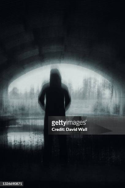 a strange hooded figure, silhouetted at the entrance to a tunnel. with a spooky, black and white blurred, vertical edit - man and his hoodie imagens e fotografias de stock
