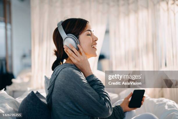 relajada joven asiática con los ojos cerrados sentada en su cama disfrutando de la música a través de los auriculares desde el teléfono inteligente en casa - headphones asian fotografías e imágenes de stock