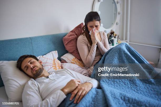 sick woman sitting on bed and blowing her nose while her husband sleeping - caucasian woman sick in bed coughing stock pictures, royalty-free photos & images