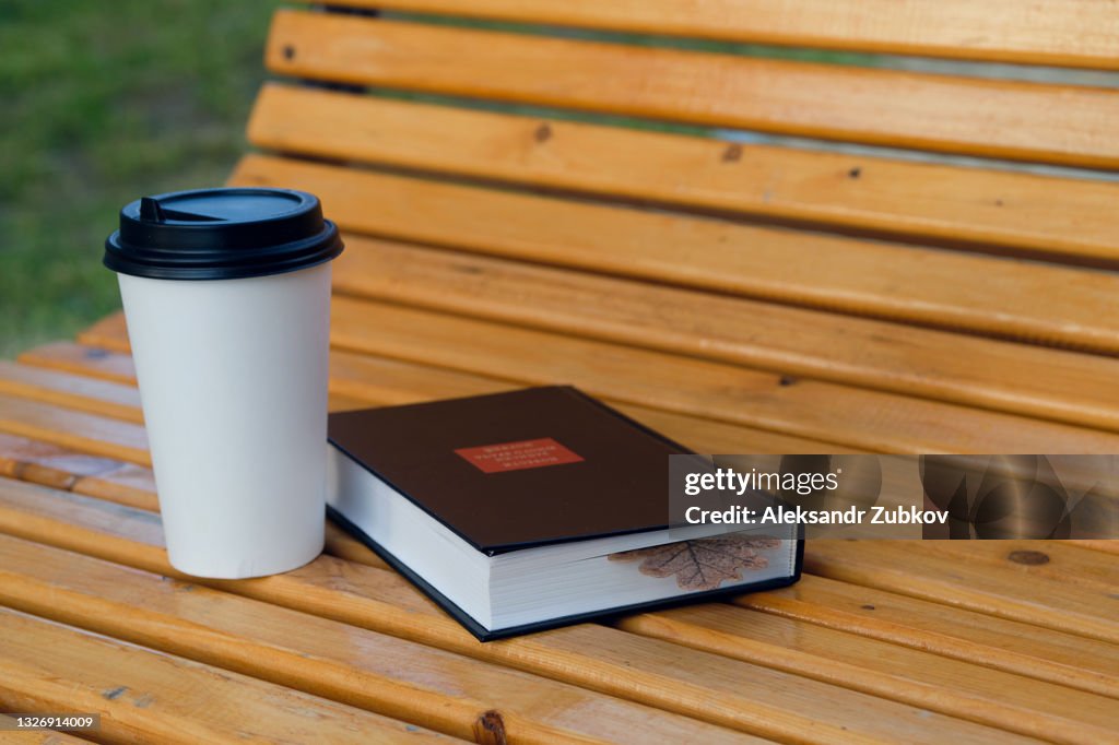 A colorful autumn oak leaf is laid between the pages of a closed book. Textbook or fiction on a wooden bench in the Park, on an autumn day. Next to it is a paper disposable Cup With Hot Warming Coffee or tea to take away.