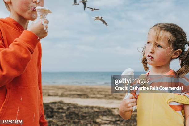two children stand on a beach by an ocean holding melting vanilla ice-creams - ice cream cone stockfoto's en -beelden