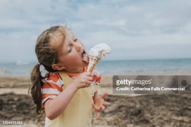 a little girl stands on a beach and eats a melting ice-cream - glace photos et images de collection