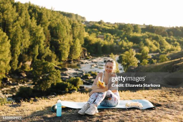 a tired girl in sports clothes is resting after a grueling sports yoga workout eating healthy healthy food fruits sitting on a sports mat in nature at the threshold of the river and the forest outdoors - banane essen stock-fotos und bilder