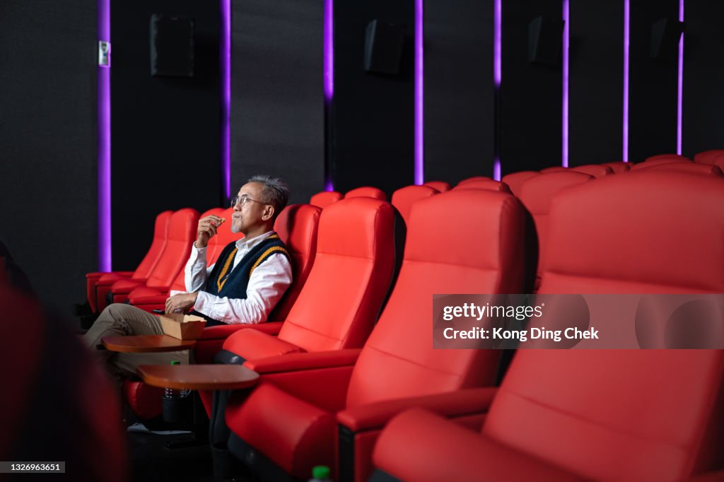 1 Asian Chinese senior man with white beard sits in empty cinema or theatre with comfortable red seats