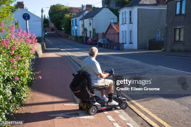 hombre en un scooter con discapacidad cede en el borde del pavimento la comprobación de la seguridad antes de conducir su scooter a través de una carretera, disfrutando de la libertad que el scooter le da para moverse por donde y cuando quiera. - mobility scooter fotografías e imágenes de stock