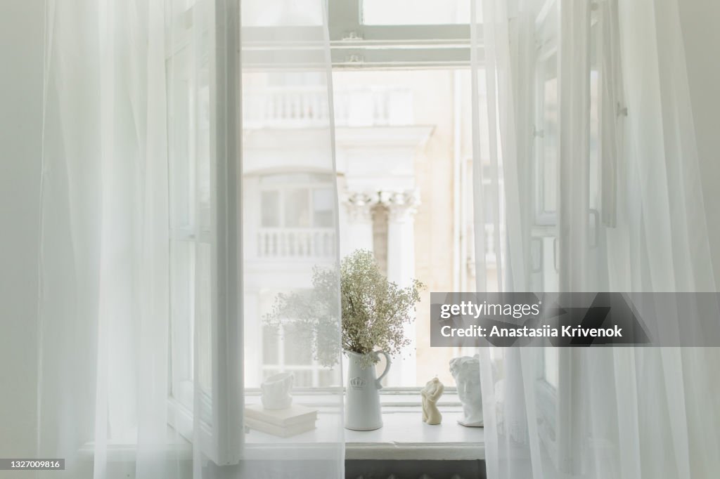 White room with curtain and and open window and vase of flowers on the windowsill.