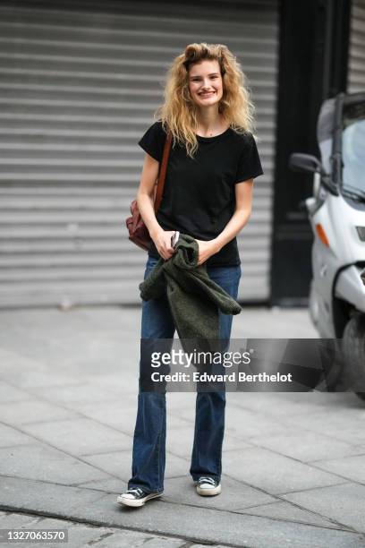 Model wears a black t-shirt, navy blue denim flared jeans pants, a dark green wool pullover, a brown shiny leather backpack bag, black Converse All...