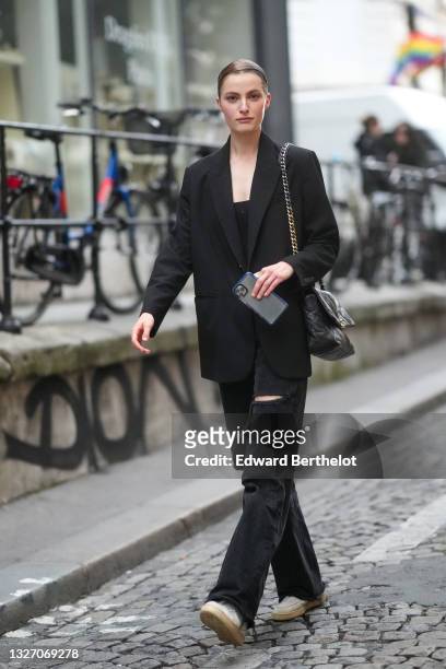 Model wears a black blazer jacket, black denim ripped jeans flared pants, a black shiny quilted leather Chanel 19 crossbody bag, a black shoulder-off...