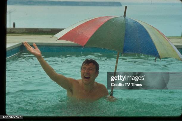 Entertainer Max Bygraves holding an umbrella in a swimming pool during a rainstorm, circa 1969.