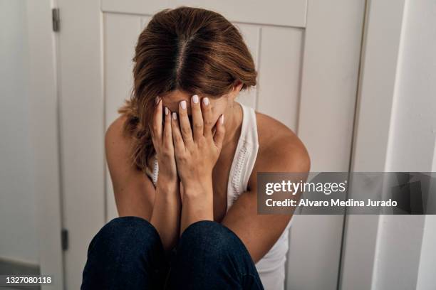unrecognizable abused woman sitting on the floor by the door, crying and covering her face with her hands. - violencia domestica imagens e fotografias de stock