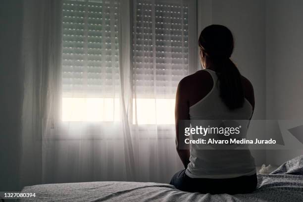 rear view of an unrecognizable abused woman sitting on her bed looking out the window. - violence stock pictures, royalty-free photos & images