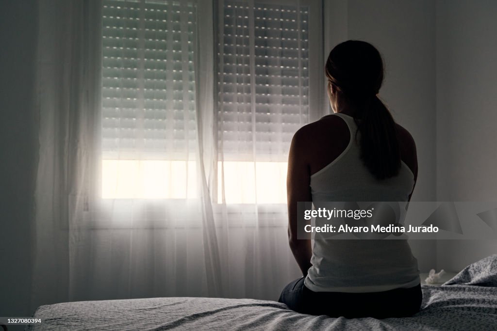 Rear view of an unrecognizable abused woman sitting on her bed looking out the window.