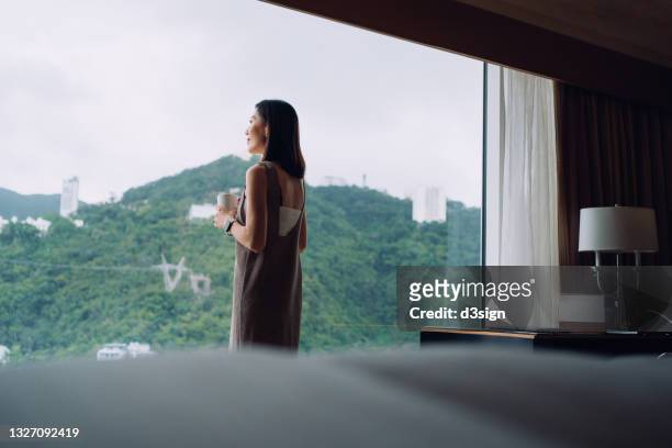 young asian woman drinking coffee, relaxing by the window at cozy home, looking out to the beautiful scenics through window in the morning . lifestyle with a moment of calm and tranquility - asian coffee at cafe imagens e fotografias de stock