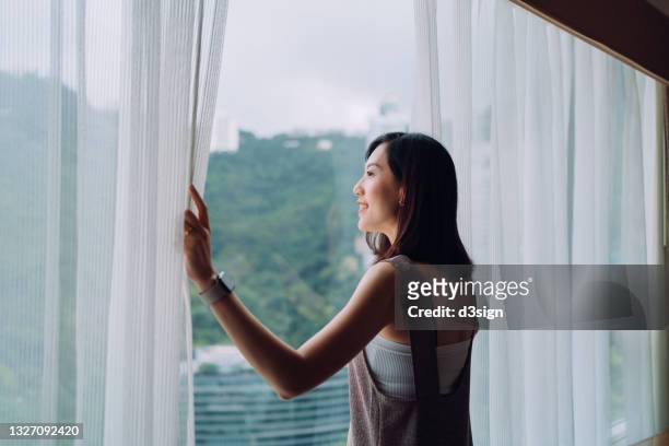 beautiful young asian woman opening curtains and looking out to the beautiful scenics through window in the morning. a brand new day with hope and possibilities - open day 3 stock pictures, royalty-free photos & images
