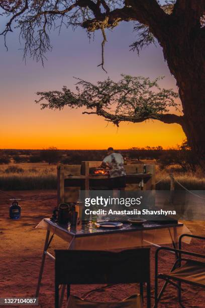 young man traveler having bbq dinner outdoors at the campsite during bright sunset in kalahari desert, namibia - kalahari desert stockfoto's en -beelden