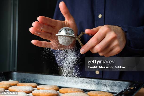 girl baker or woman pastry chef sprinkled with powdered sugar from a strainer cookies in the shape of a heart, on baking paper, on the background of a wooden kitchen table. cooking concept, valentine's day, christmas, mother's day and thanksgiving. - poedersuiker stockfoto's en -beelden