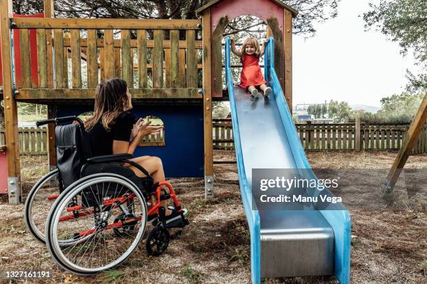 smiling mother in a wheelchair playing with her little daughter on a slide in a playground. - frau rollstuhl selbstständigkeit stock-fotos und bilder