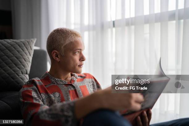 young man contemplating while reading a book at home - gay person stockfoto's en -beelden