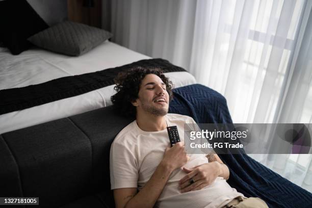 young man lying on the couch singing using remote control as microphone at home - voice remote stock pictures, royalty-free photos & images