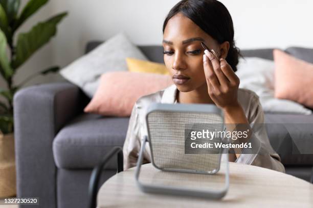 young woman using tweezers on eyebrow while looking at mirror in living room - eyebrow tweezers imagens e fotografias de stock