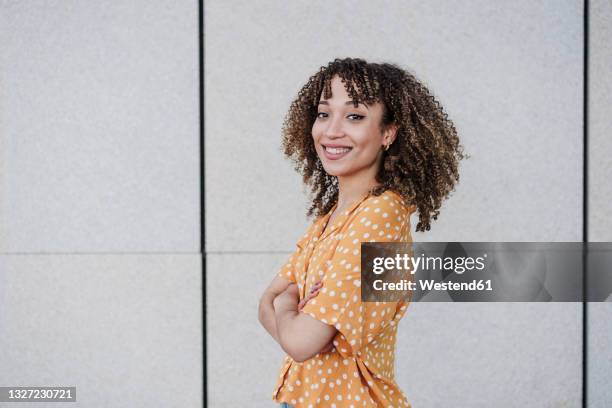 smiling young woman with arms crossed standing by wall - curly bangs stock pictures, royalty-free photos & images