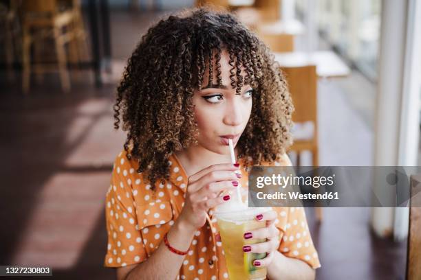 young woman with curly hair looking away while drinking iced tea in cafe - ice tea stock-fotos und bilder