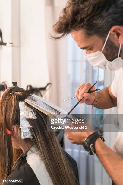 male hairdresser with protective face mask dyeing hair at salon - dye imagens e fotografias de stock