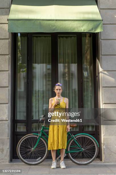 woman holding smoothie while standing with bicycle in the city - yellow smoothie stock pictures, royalty-free photos & images