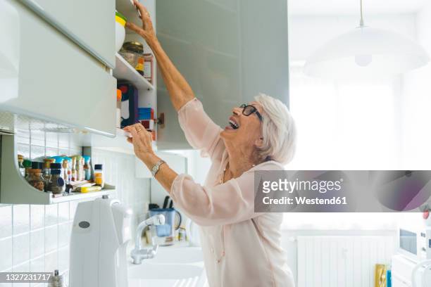 cheerful senior woman doing chores in kitchen at home - cabinet stock pictures, royalty-free photos & images