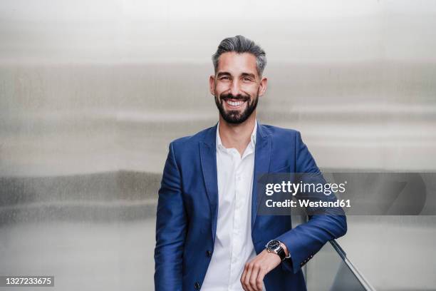 mid adult businessman smiling while leaning on railing in front of silver colored wall - veste homme photos et images de collection