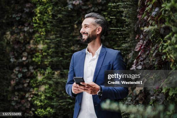 male entrepreneur with mobile phone smiling while standing by plants - living_walls stock pictures, royalty-free photos & images