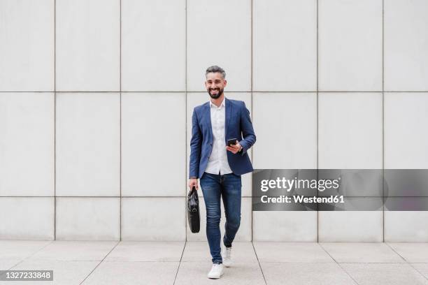 smiling businessman with briefcase and mobile phone walking on footpath - mann frontal ganzkörper jacket stock-fotos und bilder