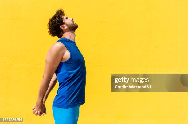 mid adult man doing stretching exercise by yellow wall during sunny day - mani dietro la schiena foto e immagini stock