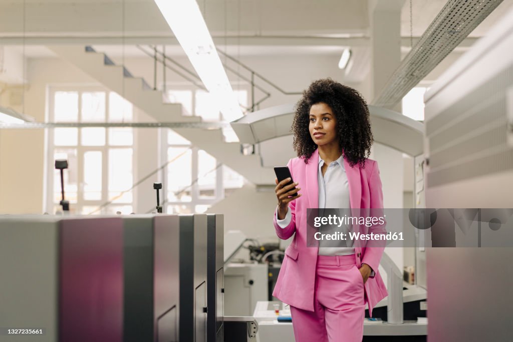 Female professional with mobile phone looking away while standing amidst machines in industry